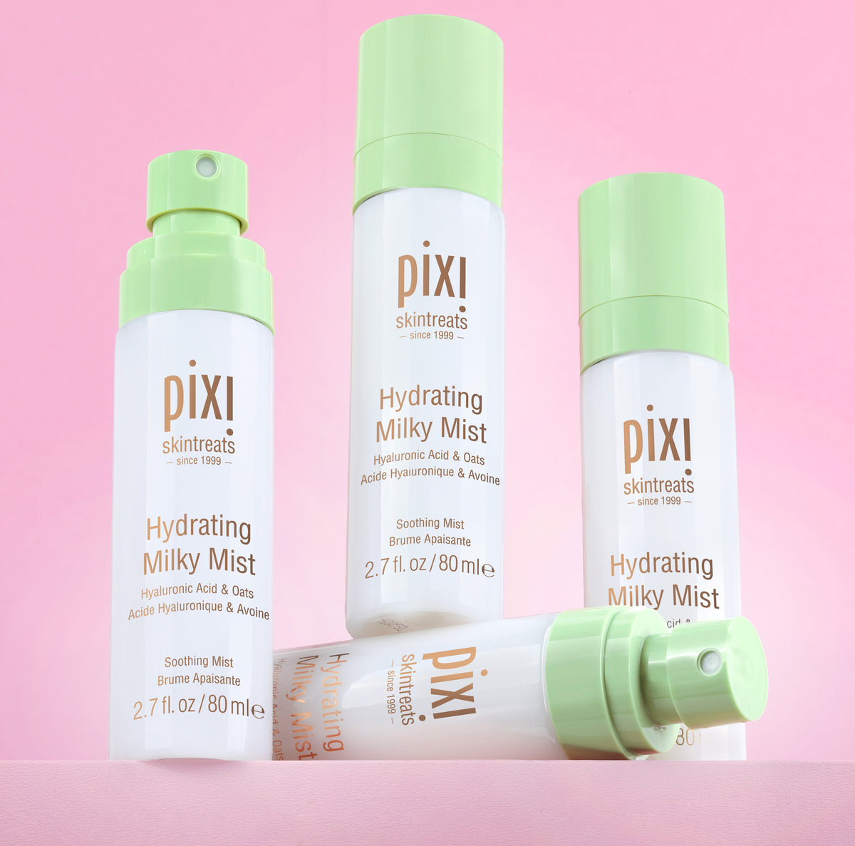 Three Pixi Skintreats Hydrating Milky Mist bottles in white and light green packaging, arranged on a solid pink background with one bottle lying down in front.