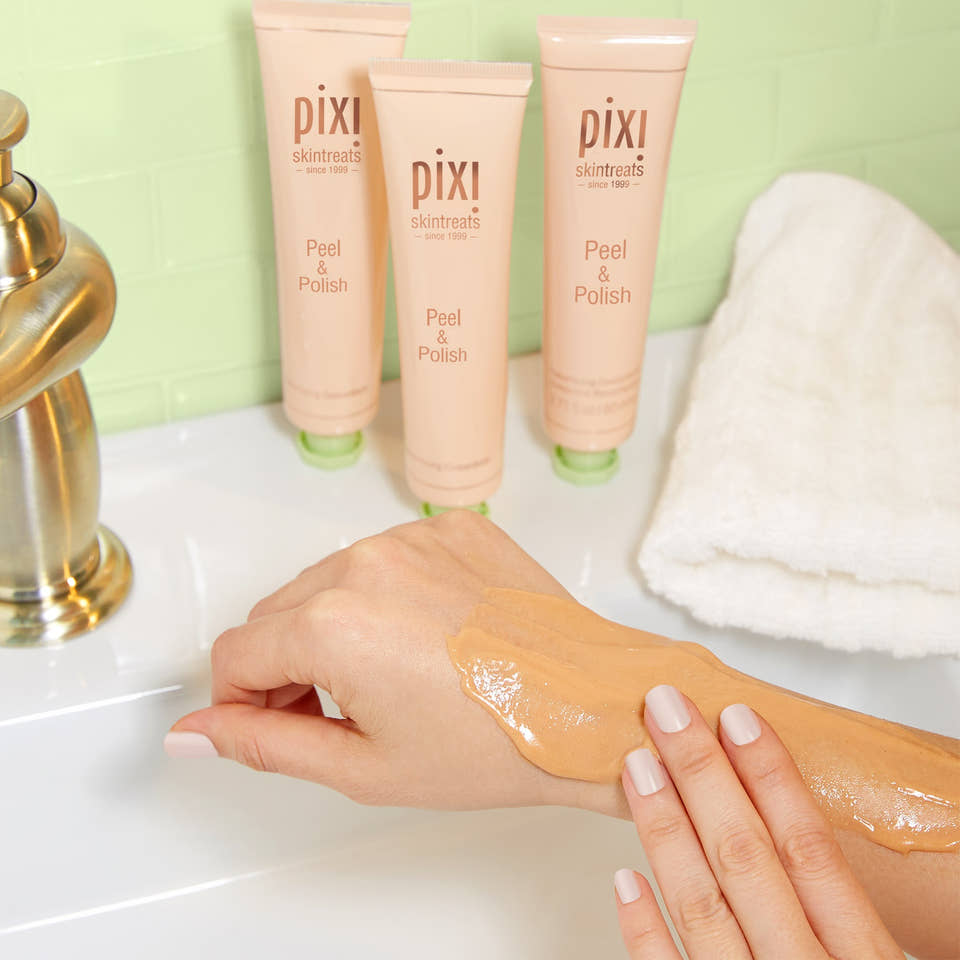 Three tubes of pixi skin treats Peel & Polish cream are lined up on a bathroom counter next to a folded white towel, with a hand showing the thick, glossy cream product applied to the forearm. view 2